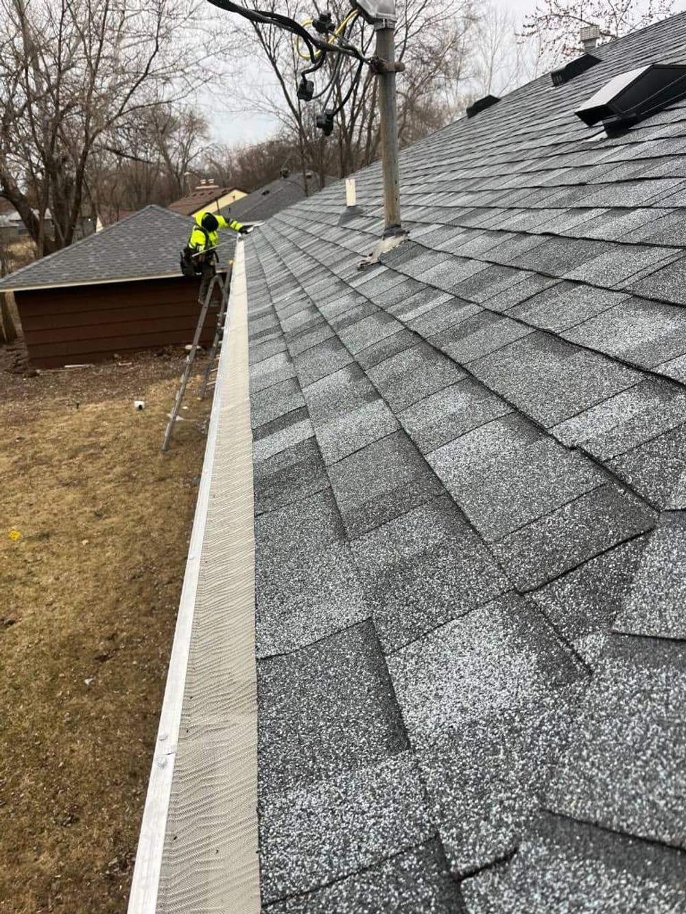 Roof inspection with worker on ladder, showcasing shingles and nearby trees in winter setting.