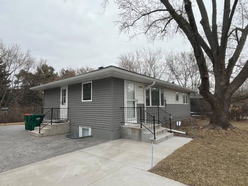 Gray bungalow home with a landscaped front yard and concrete driveway, featuring green garbage bins.