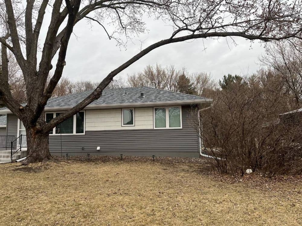 Gray house exterior with large tree and overcast sky, surrounded by brown grass and shrubs.