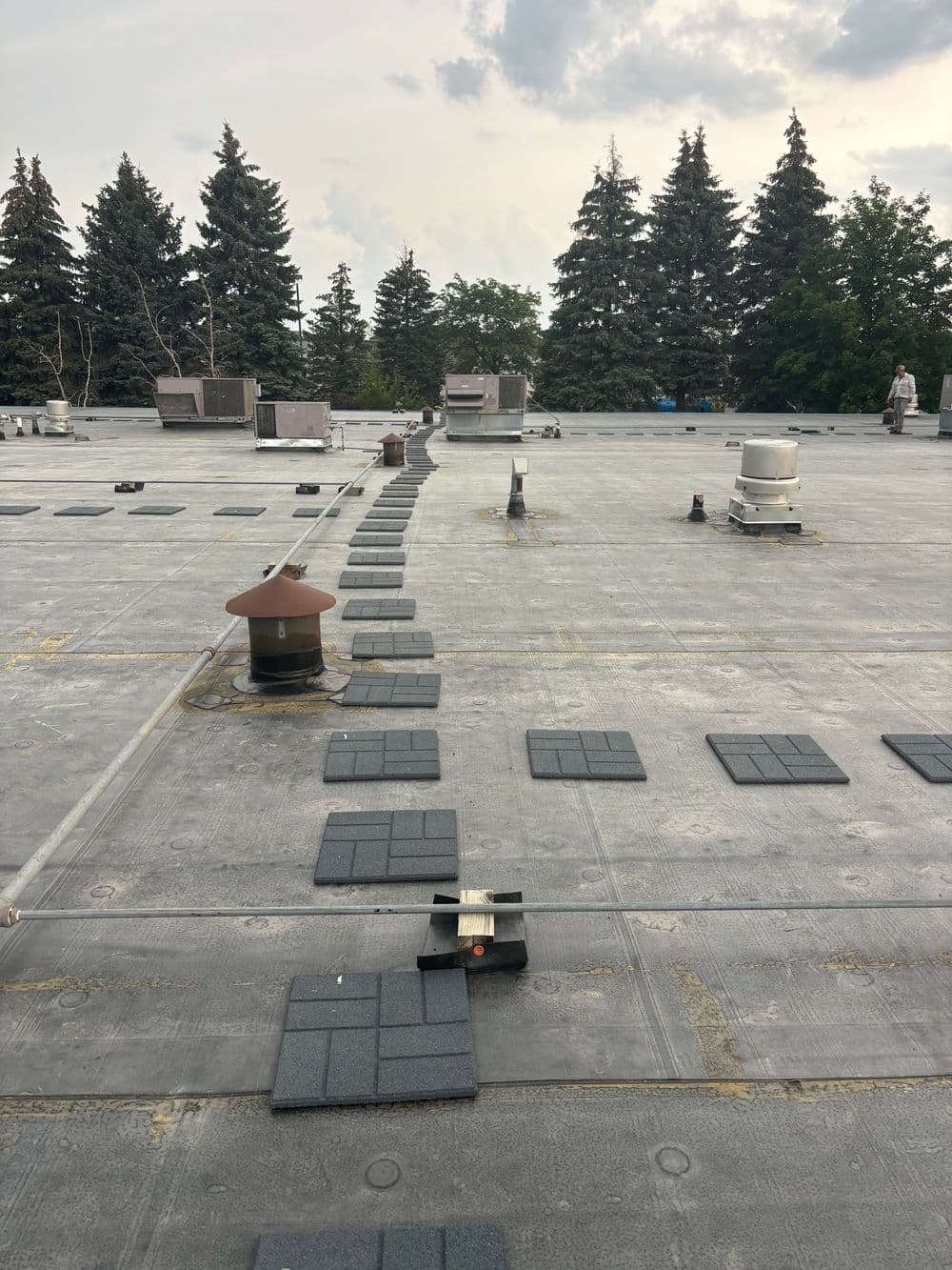 Flat commercial rooftop with HVAC units and gray pavers under a cloudy sky. Trees in the background.