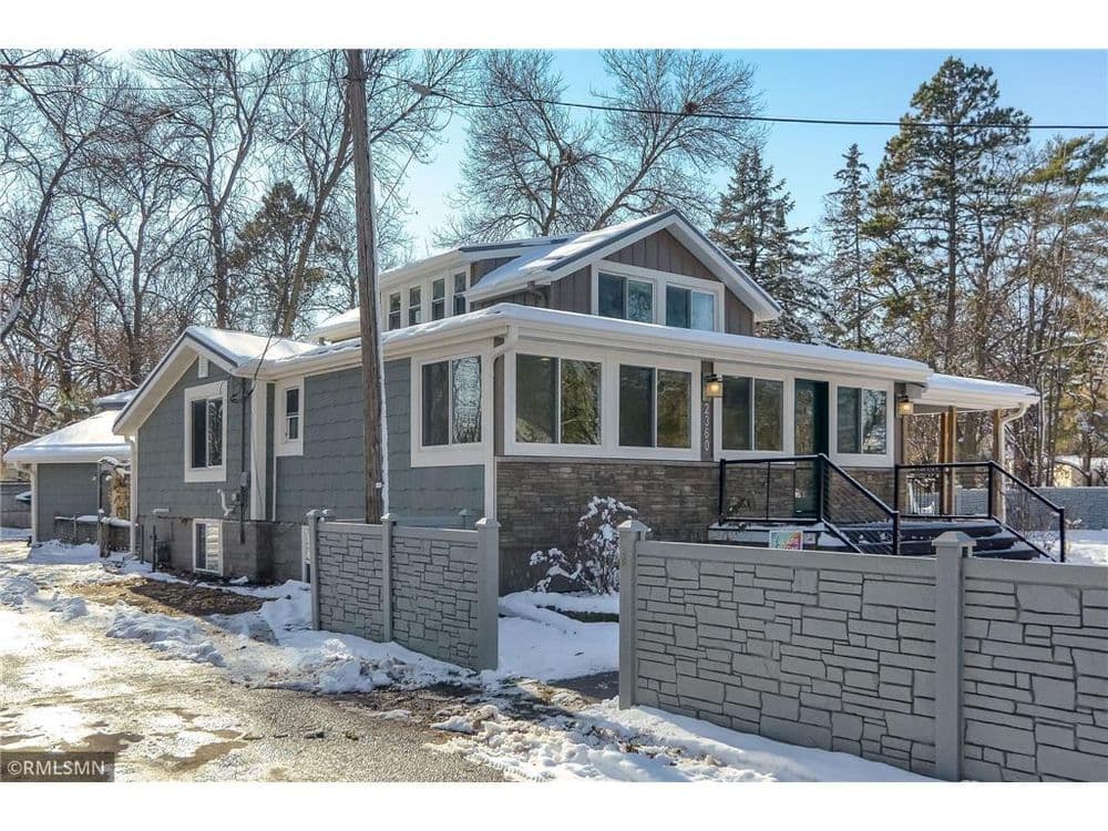 Two-story gray house with large windows and stone accents, surrounded by snow and trees.