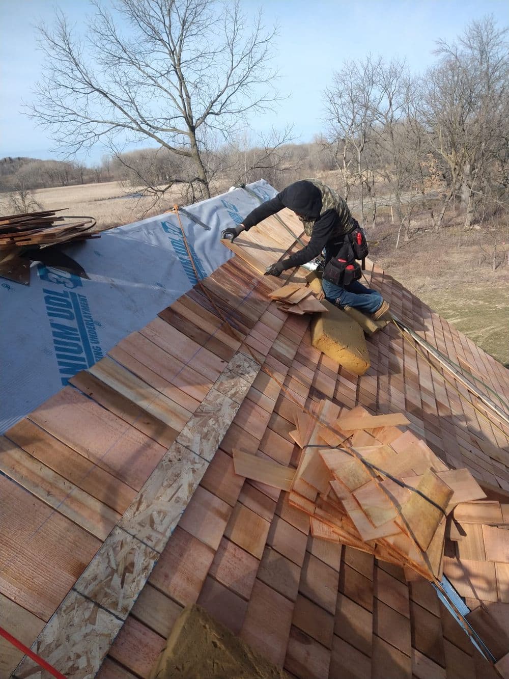 Roofer installing cedar shingles on a steep roof in a rural landscape. Safety harness visible.