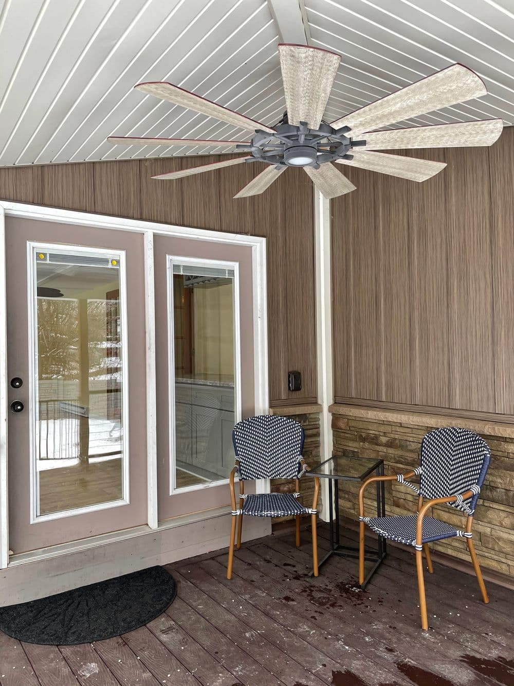Modern porch with a ceiling fan, two patterned chairs, and a glass table beside double doors.