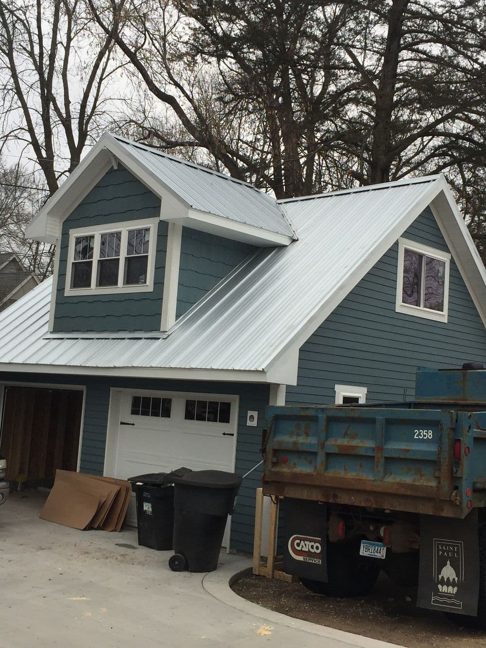 Blue house with metal roof and garage, surrounded by trees. Trash bins in front.
