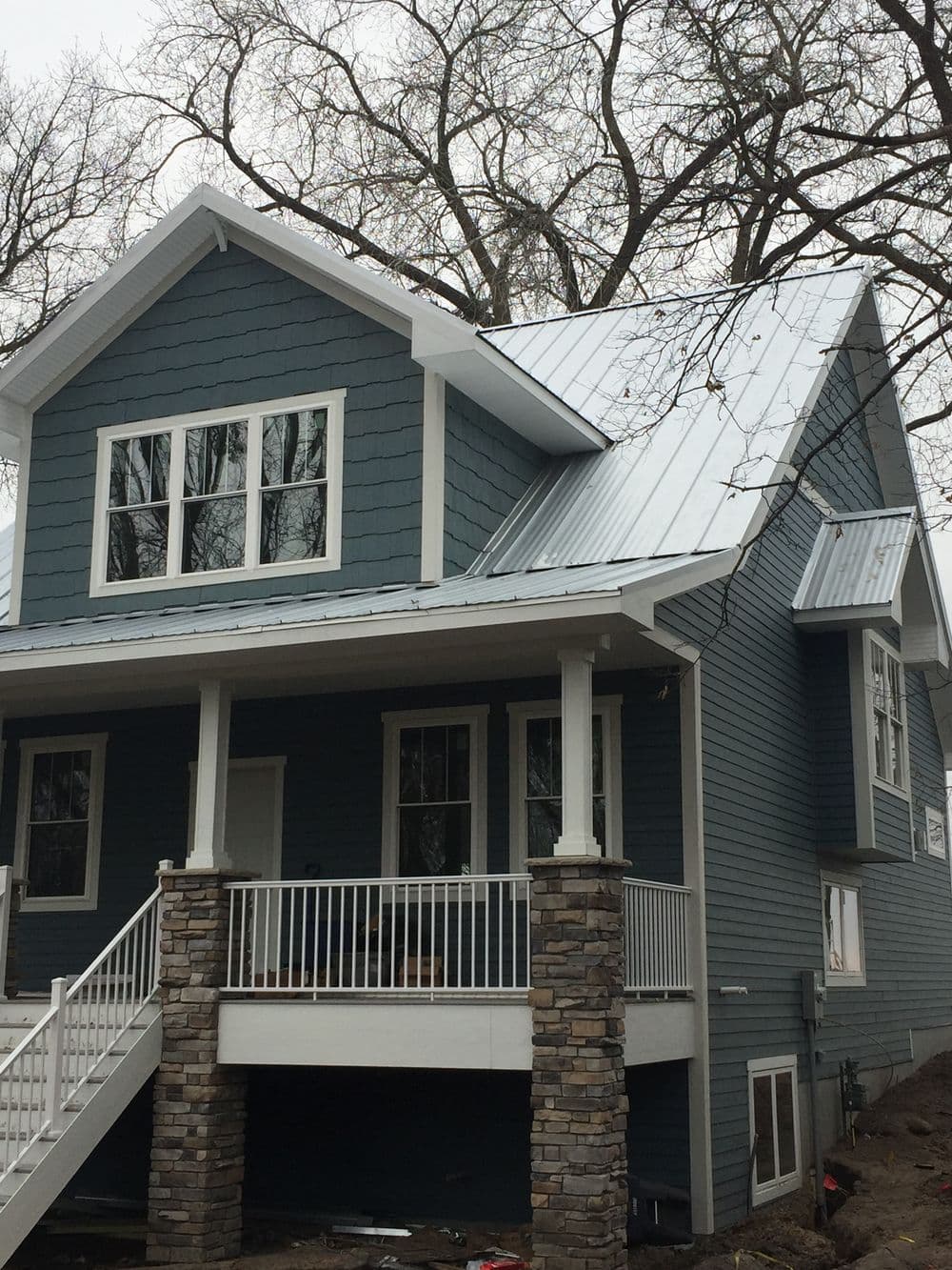 Modern blue house with a metal roof, stone columns, and large windows in a wooded area.