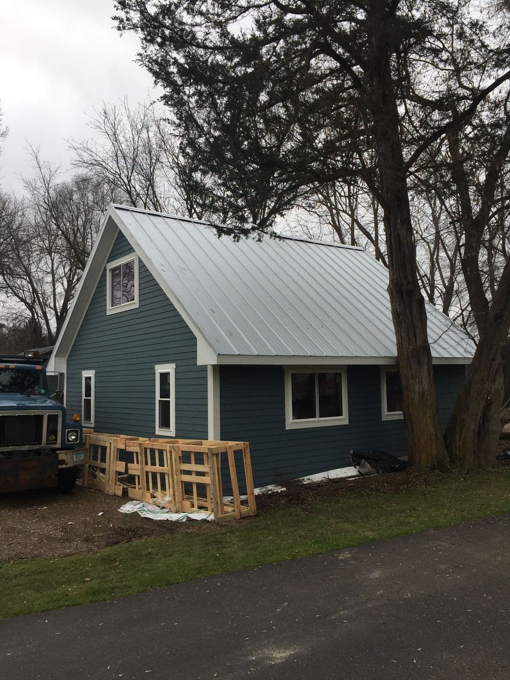 Modern blue house with a metal roof surrounded by trees and construction materials.